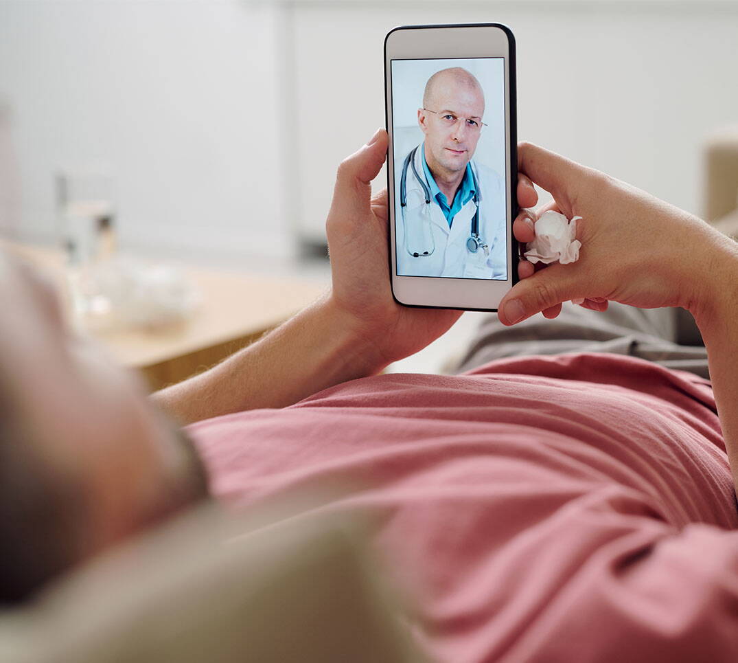 Kranker Mann liegt auf dem Sofa und lässt sich per Video-Anruf auf dem Smartphone von einem Arzt beraten