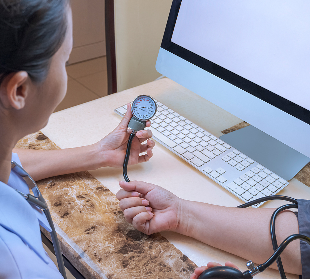 Santé-numérique-Suisse-TARDOC-2026 Médecin prenant la tension artérielle sur un lieu de travail équipé d’un ordinateur – numérisation du système de santé suisse grâce à la réforme TARDOC 2026