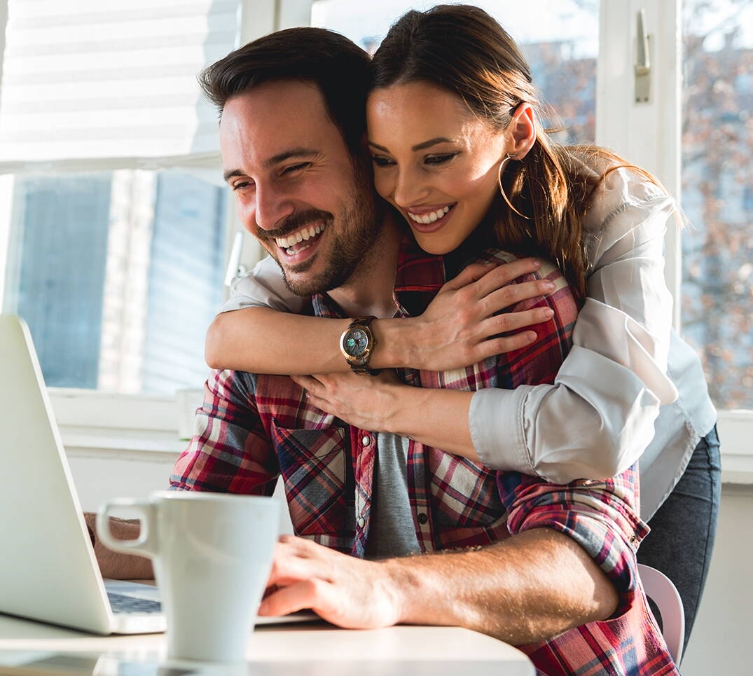 Una giovane coppia sorridente seduta davanti a un computer portatile si informa su come risparmiare sui premi dell’assicurazione malattia.