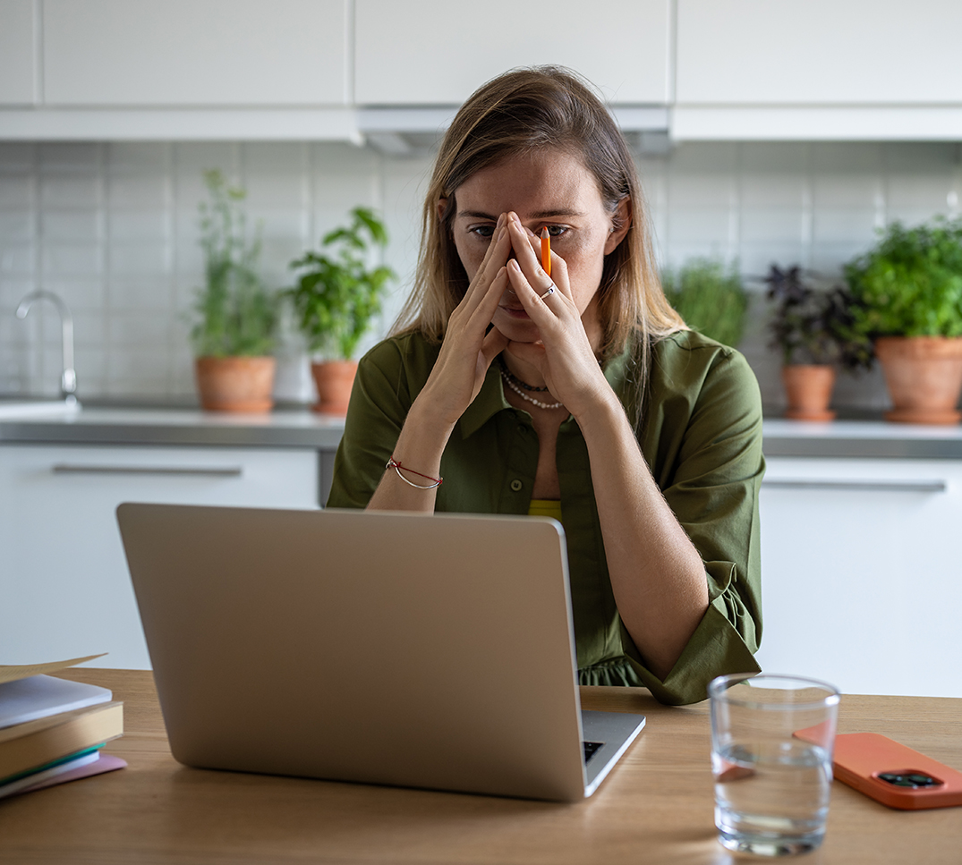 Une personne est assise à une table dans une cuisine et travaille sur un ordinateur portable. Des documents, un verre d’eau et du matériel d’écriture se trouvent sur la table; des plantes d’intérieur sont visibles à l’arrière‑plan.