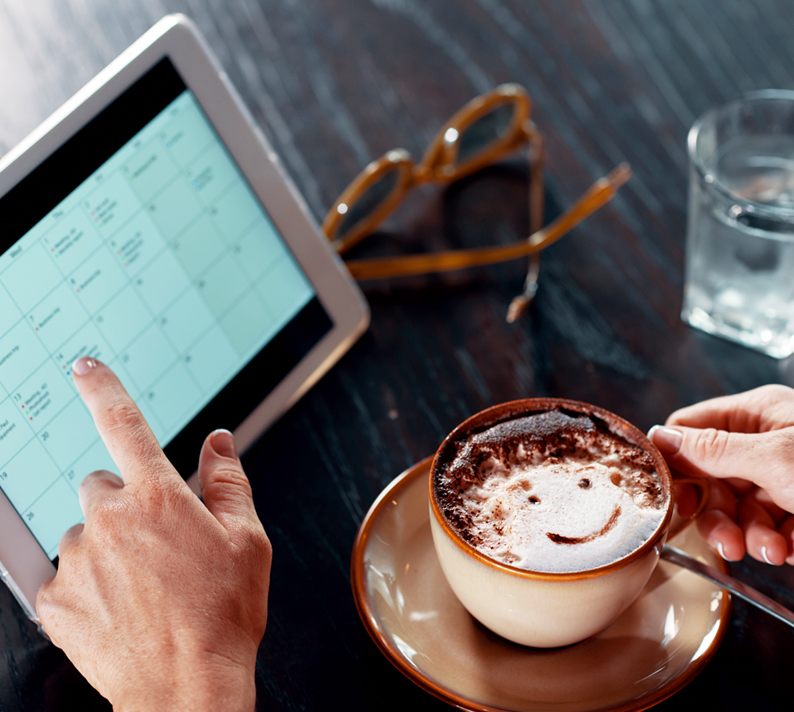 Eine Hand zeigt auf ein Tablet mit einem Kalender. Daneben stehen eine Brille, ein Glas Wasser und eine Tasse mit einem Muster auf dem Milchschaum.