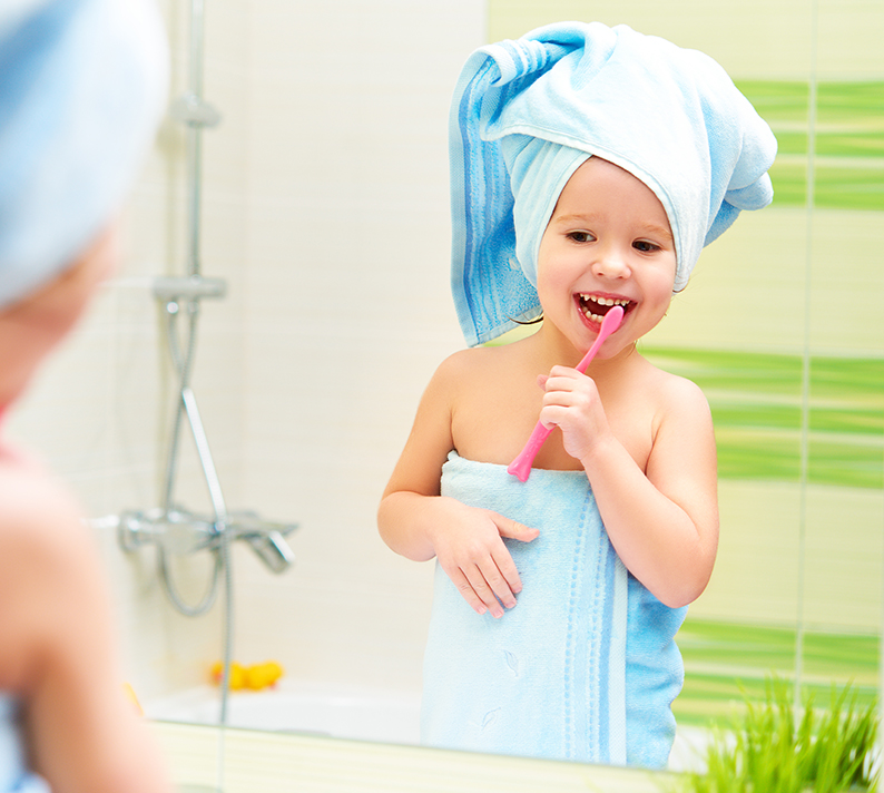 Un bambino è davanti allo specchio del bagno, avvolto in un asciugamano e con uno spazzolino da denti in mano. Sullo sfondo si vedono la vasca da bagno e piastrelle verdi.